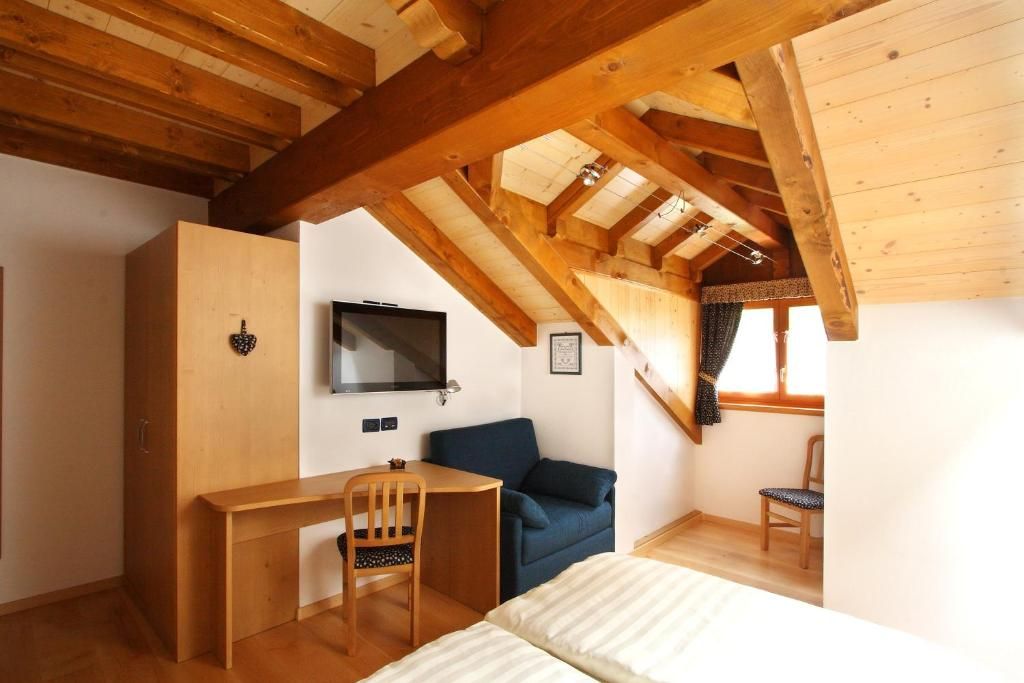 A room with a wooden ceiling, a bed, a desk with a chair, a television, and a window with curtains.