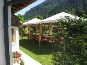 A covered outdoor dining area with tables and chairs, surrounded by greenery and a mountainous backdrop.