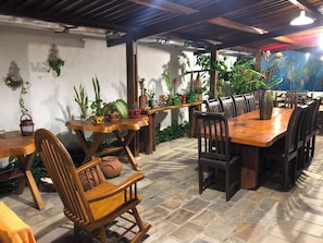 Uma sala de jantar com mesas e cadeiras de madeira, plantas em vasos e teto rústico.