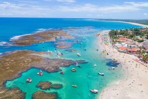 Vista aérea de una zona costera con aguas turquesas cristalinas, barcos y una playa con sombrillas y gente.