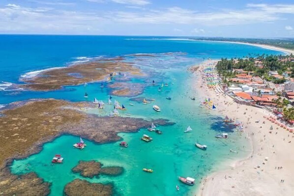 Vista aérea de uma área costeira com águas turquesa cristalinas, barcos e uma praia com guarda-sóis e pessoas.