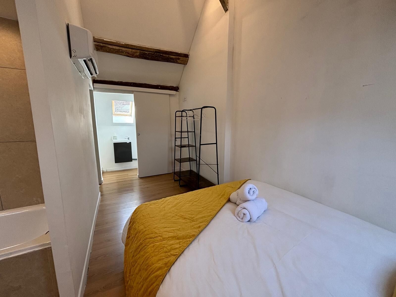 Une petite pièce étroite, avec un lit, une couverture jaune et une serviette blanche. Contre le mur, on trouve une étagère en métal noir, et une porte donne accès à une autre pièce.