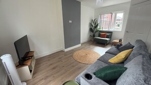 Una sala de estar con un sofá gris, una mesita pequeña verde, un ventilador de pie, un televisor sobre un mueble de madera, una planta en maceta y una ventana con vistas a edificios.