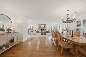 Uma sala de estar ampla com lareira, uma área de jantar com mobiliário de madeira e uma cozinha ao fundo.