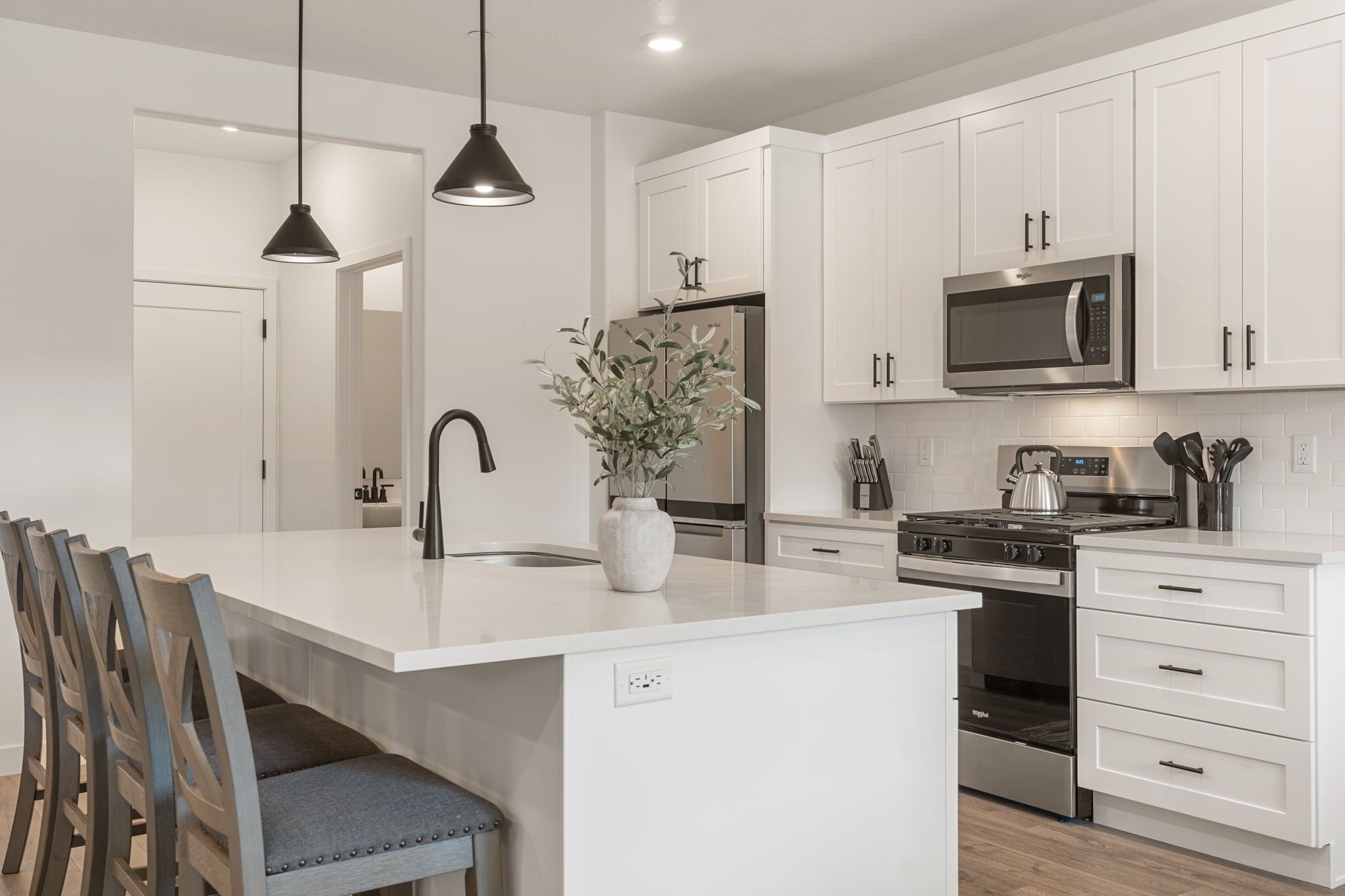 A modern kitchen with white cabinetry, stainless steel appliances, a central island with seating, pendant lighting, and a potted plant.