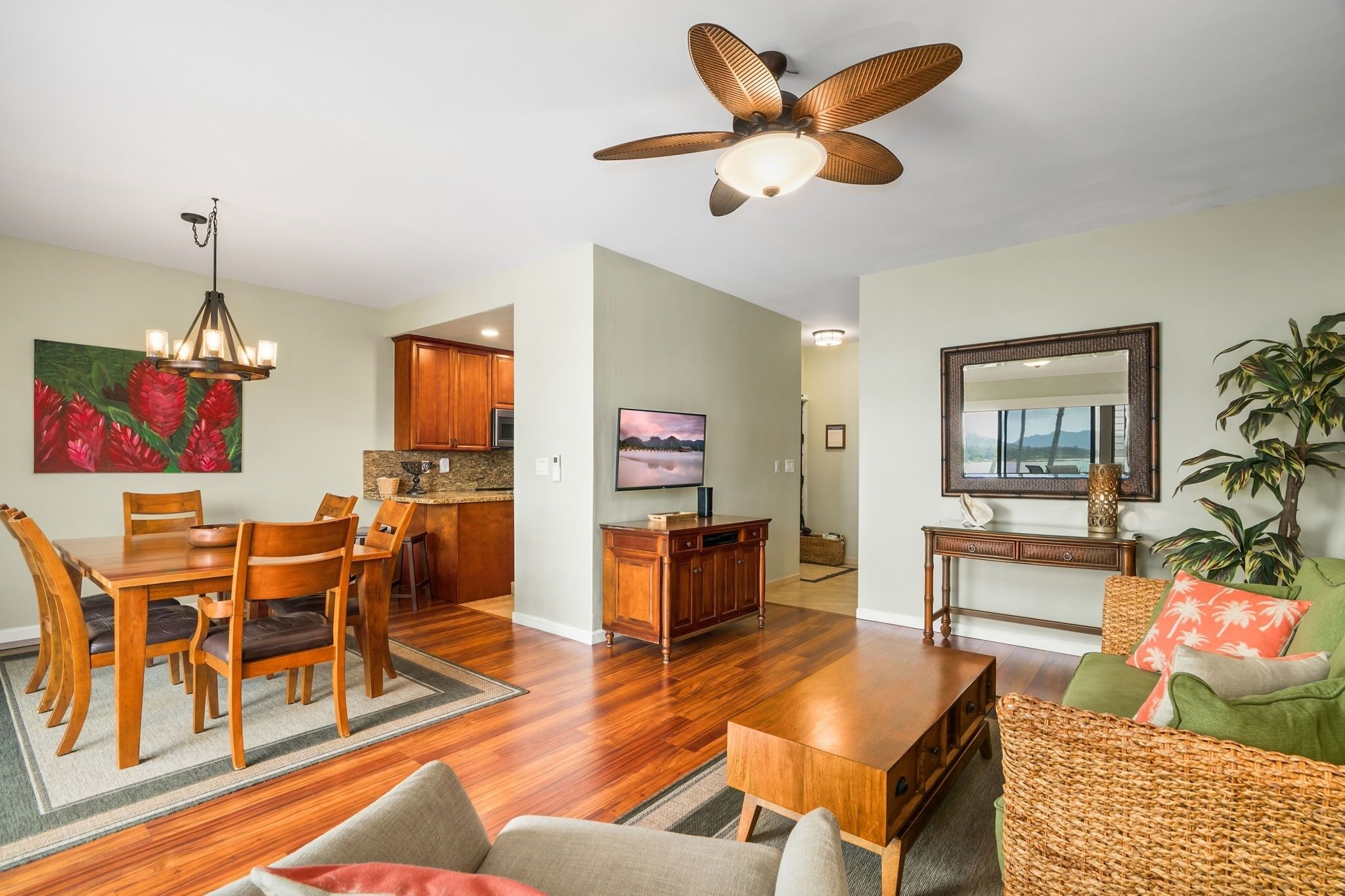 A living room with a dining table, chairs, a sofa, a TV, a ceiling fan, and a kitchen in the background.
