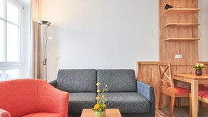 Ett vardagsrum med en grå soffa, en röd fåtölj, ett träbord med en vas med blommor, ett matbord med stolar och en hyllsektion i trä.
