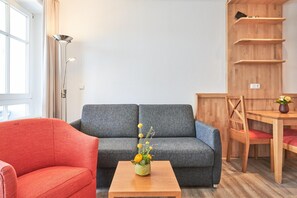 Ein Wohnzimmer mit grauem Sofa, einem roten Sessel, einem Holztisch mit Blumenvase, einem Esstisch mit Stühlen und einer Holzregaleinheit.