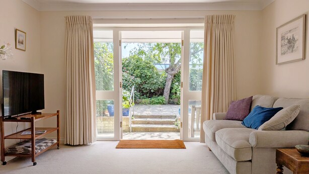 A living room with a television on a wooden stand, a beige sofa with cushions, a wooden side table, and a view through a glass door to an outdoor area with steps and greenery. - Apple Cottage - Emsworth  -  a cottage that sleeps 5 guests  in 3 bedrooms (Emsworth)
