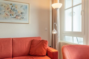 A room with a red sofa, a floor lamp, a window with curtains, and a framed artwork on the wall. - Ferienwohnung Sellin auf Rugen mit Balkon (Sellin)