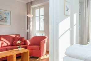 A room with a red sofa, a wooden coffee table, a white bed, and a large window with curtains. - Ferienwohnung Sellin auf Rugen mit Balkon (Sellin)