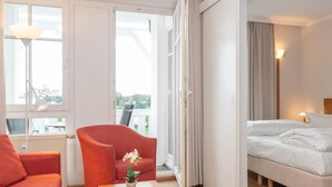 A hotel room with a bed, a red armchair, a small table with a potted plant, and a view of the outdoors through large windows. - Ferienwohnung Sellin auf Rugen mit Balkon (Sellin)