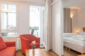 Ein Hotelzimmer mit einem Bett, einem roten Sessel, einem kleinen Tisch mit Topfpflanze und Blick ins Freie durch große Fenster.
