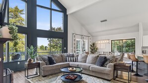 A modern living room with a sectional sofa, a coffee table, and large windows. - Fairway to Heaven: A-frame Retreat with Hot Tub, Arcade, Golf and More. Dogs OK. (Hedgesville)