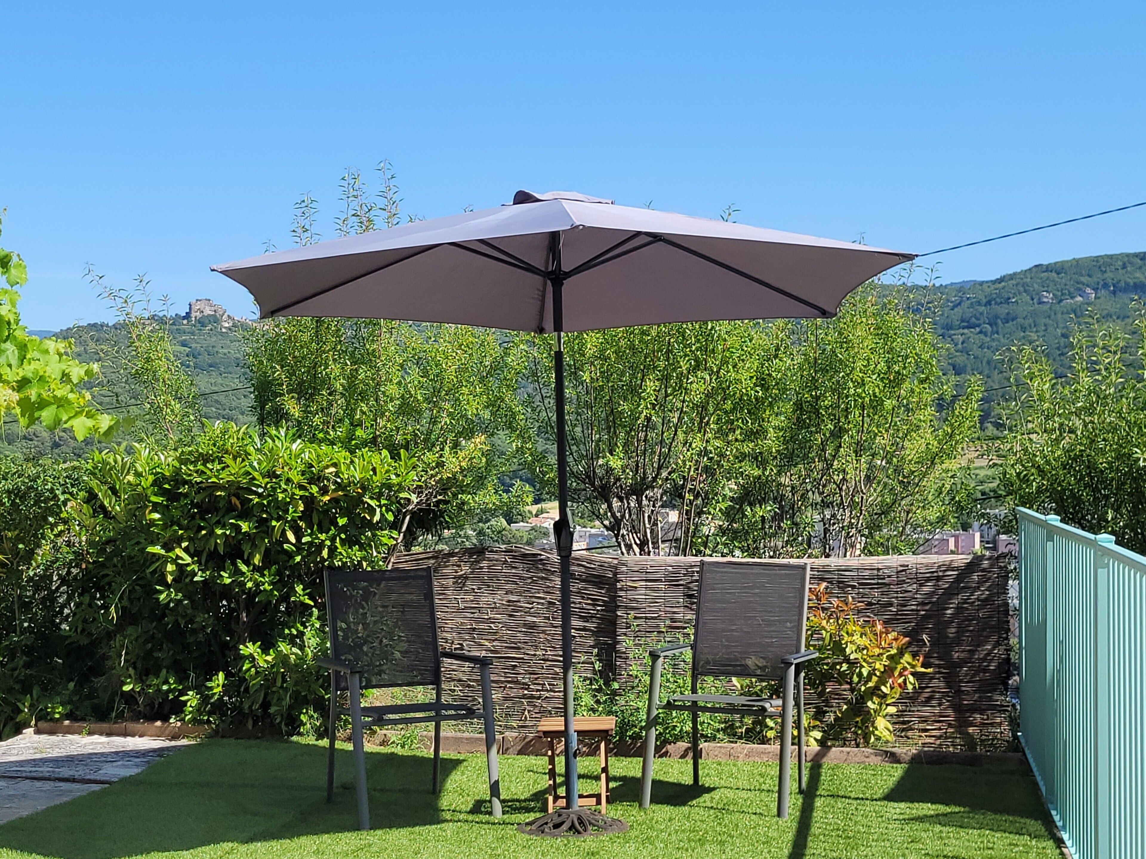 Une terrasse avec une table et des chaises, un grand parasol et une vue sur une colline bordée d’arbres.
