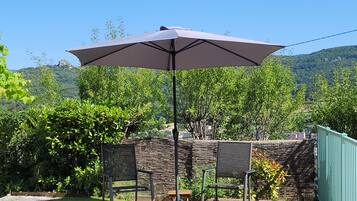 Une terrasse avec une table et des chaises, un grand parasol et une vue sur une colline bordée d’arbres.