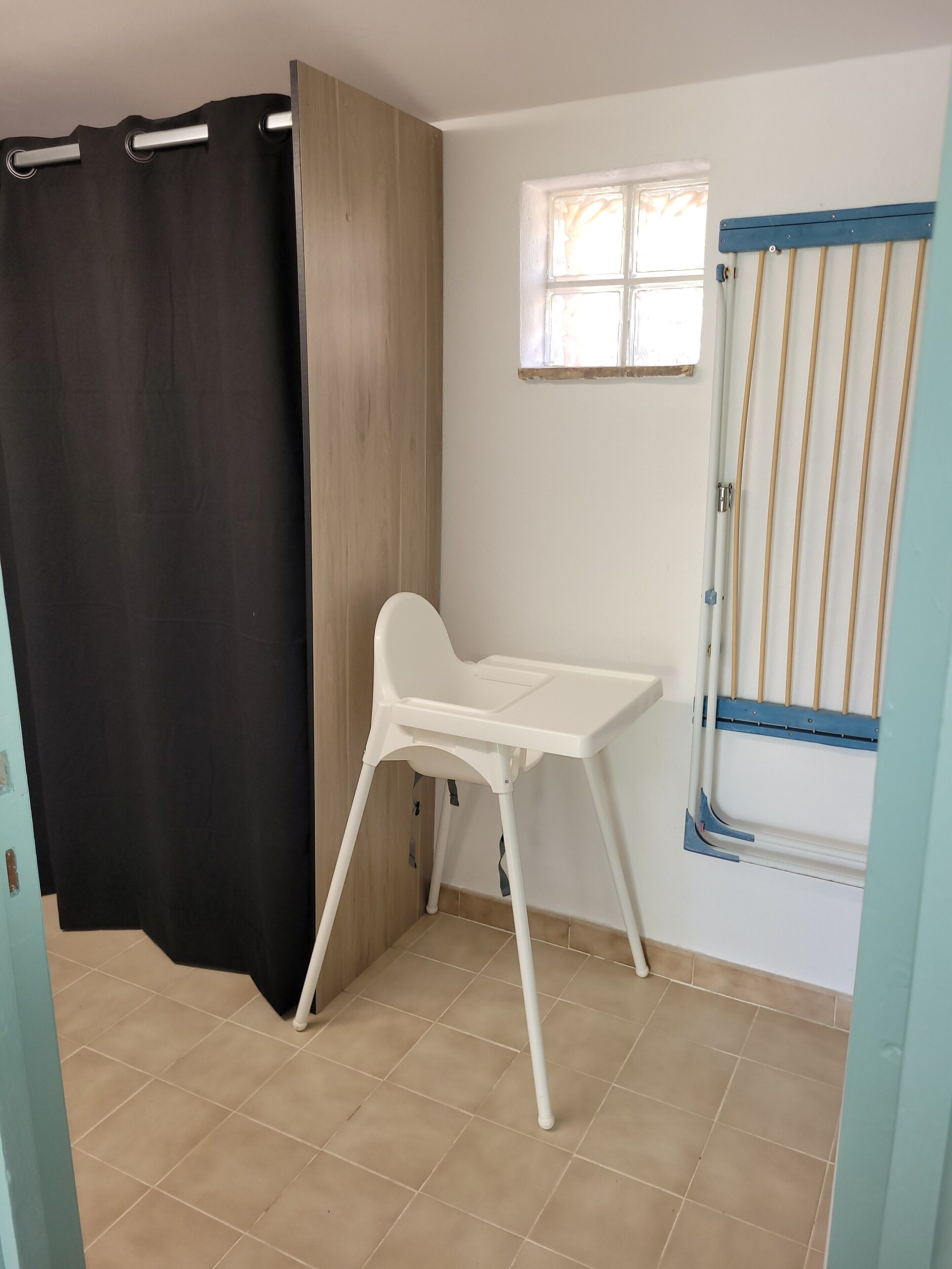 A room with a white baby changing table, a black curtain, a wooden door, a window with blinds, and a blue-framed window blind.