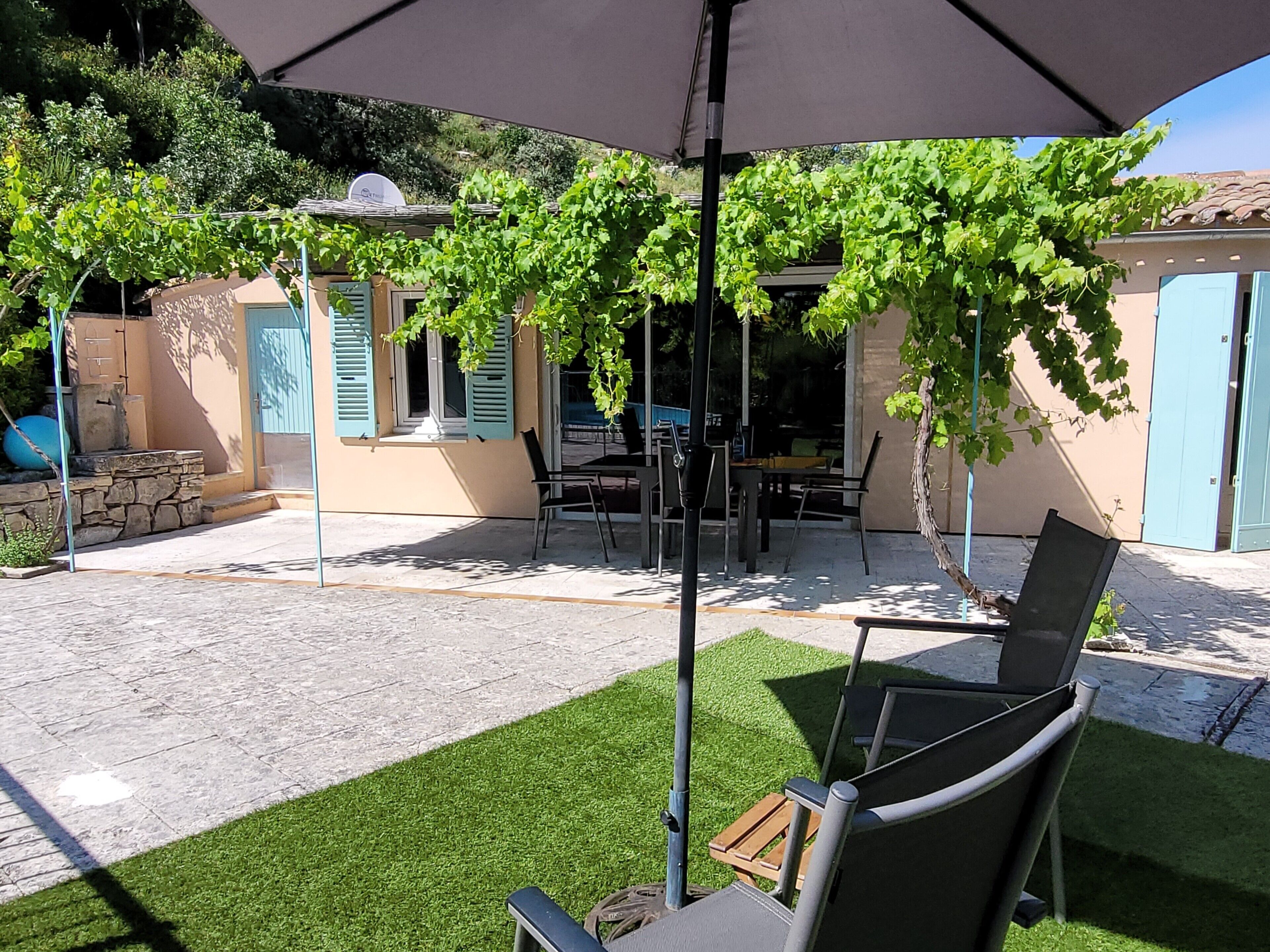 Une terrasse extérieure avec une table et des chaises, une pergola couverte de plantes grimpantes et un bâtiment aux volets bleus.