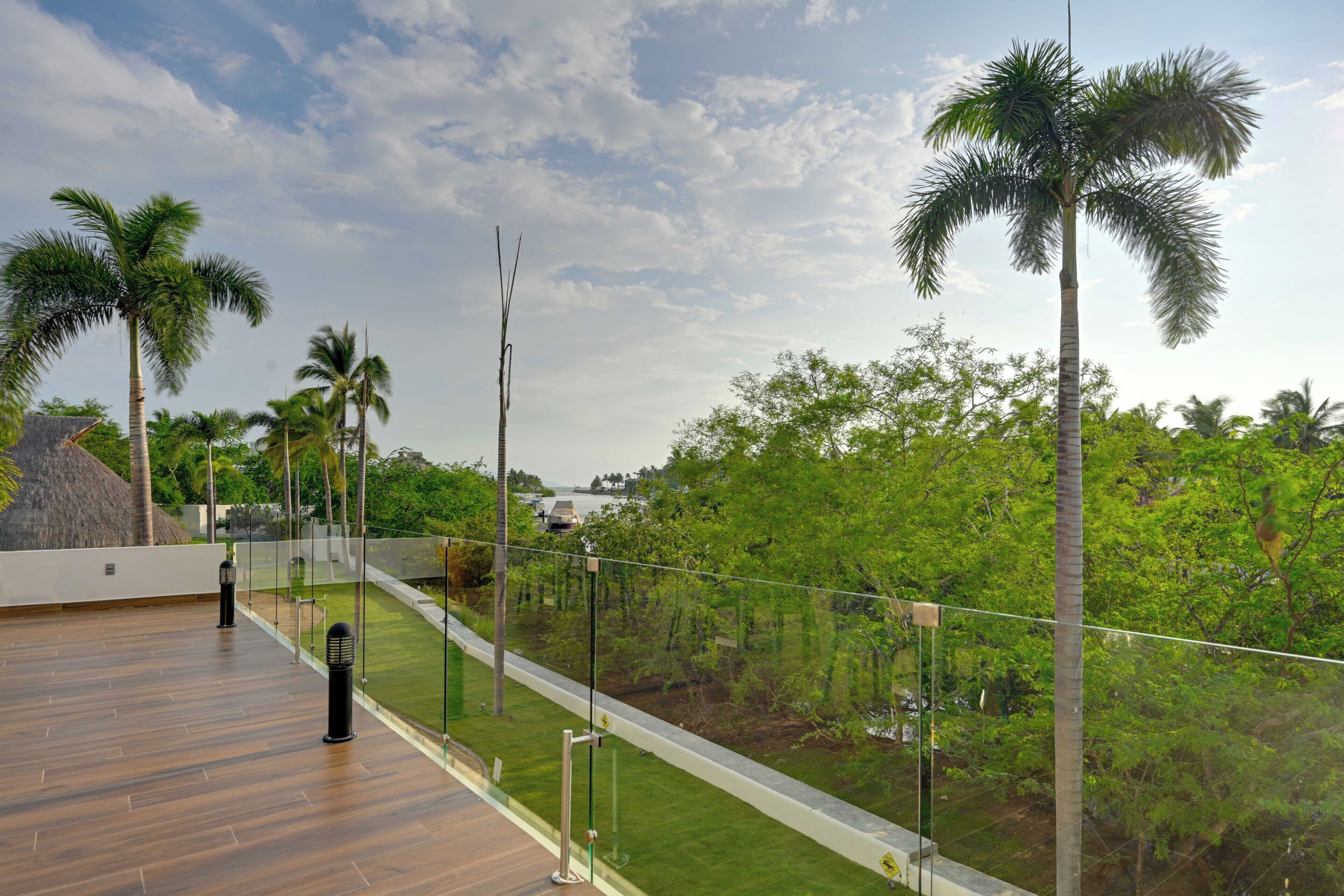 A wooden deck with a glass railing overlooking a lush green landscape with palm trees.