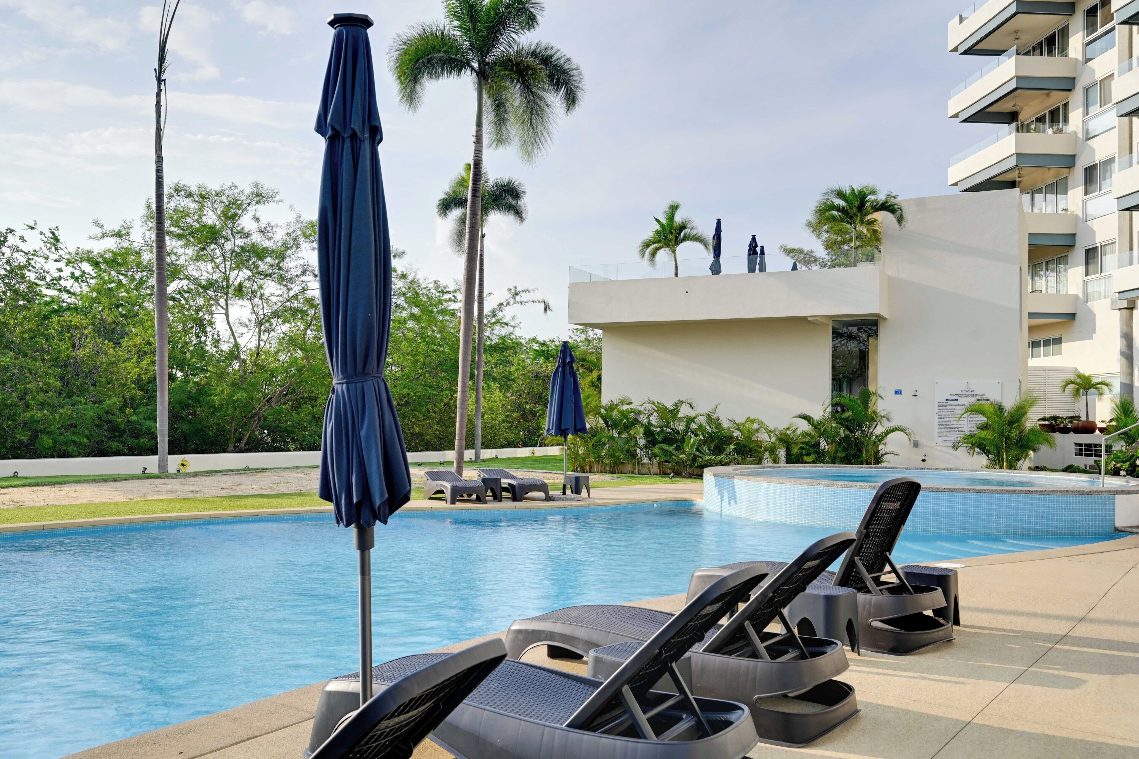 Una piscina con sillones de descanso y sombrillas, rodeada de palmeras y un edificio moderno.