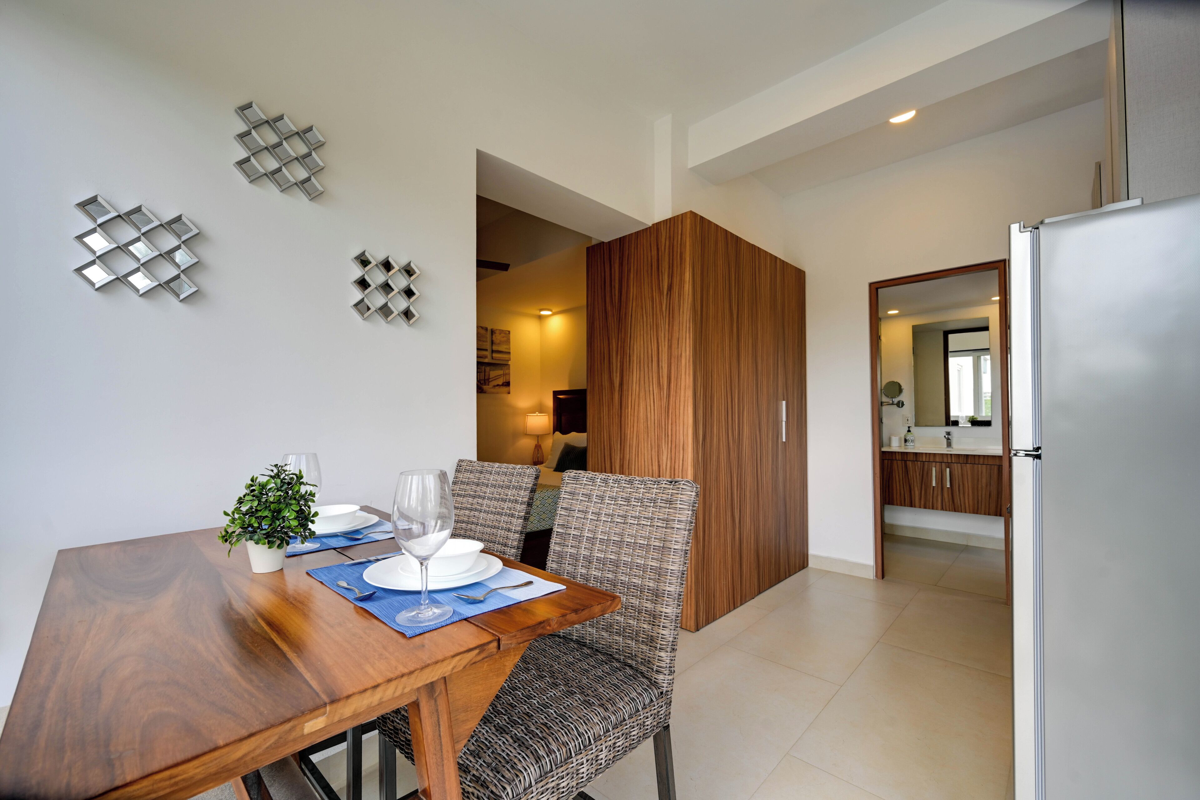 Un comedor con una mesa de madera, sillas y una planta pequeña en maceta. Hay decoraciones en la pared y se ve entrada a otra habitación.