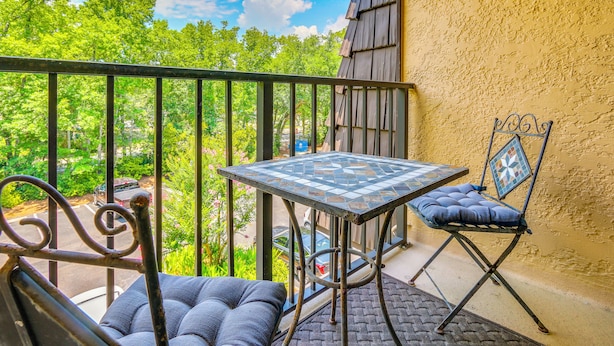 A balcony with a table and chairs, a view of trees, and a building with a textured wall. - Pool & Beach! Lagoon-view Island Club Resort Condo (Hilton Head Island)