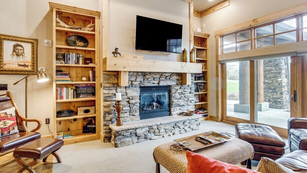 A cozy living room with a stone fireplace, wooden bookshelves, leather furniture, and a large window. - Gateway to the Teton Valley! Luxe Mtn Condo (Victor)