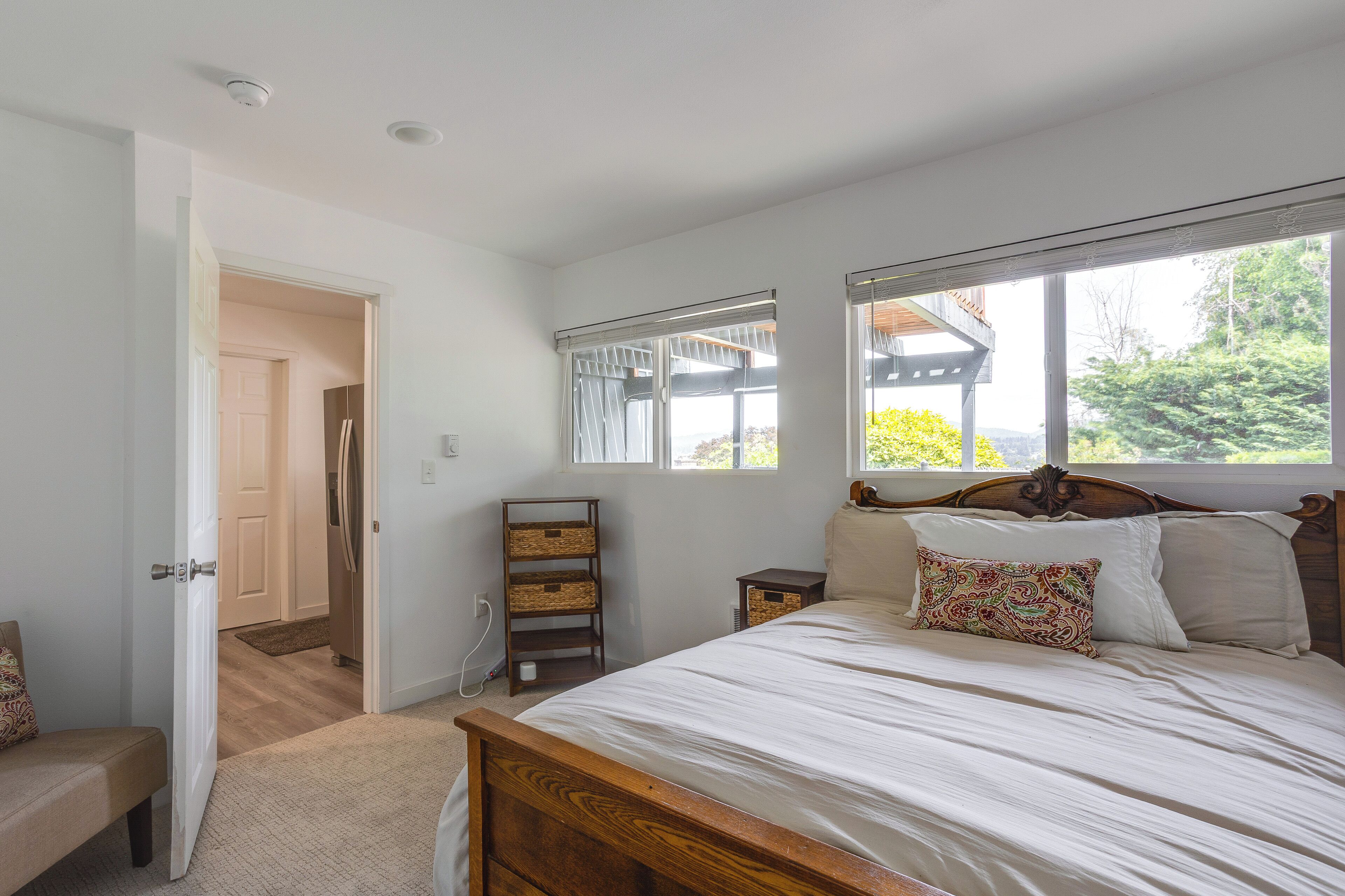 A bedroom with a wooden bed, a small bedside table, a nightstand, and a window with a view of greenery.