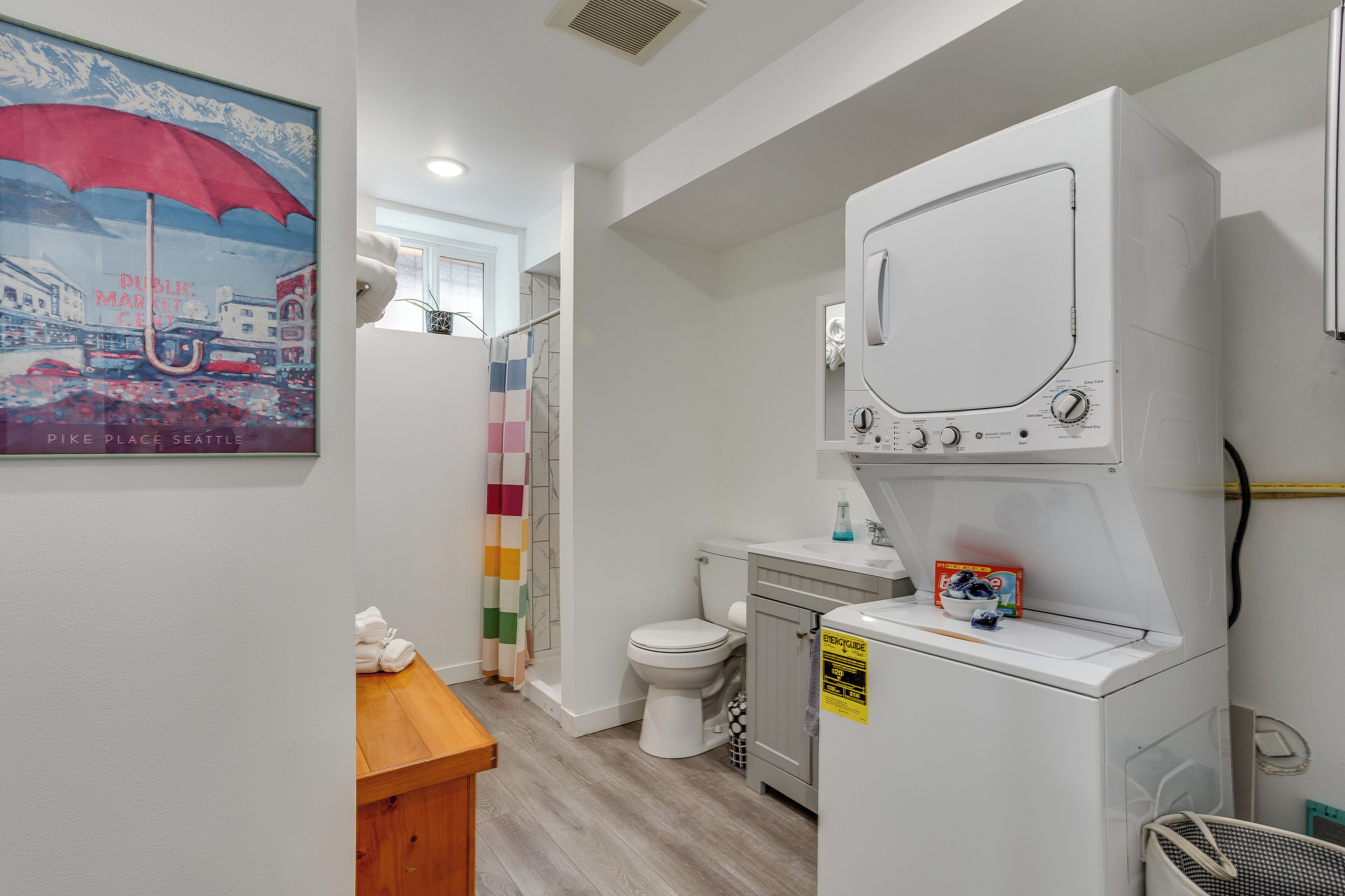 A compact hotel bathroom with a white washing machine, a toilet, a wooden bench, and a colorful shower curtain.
