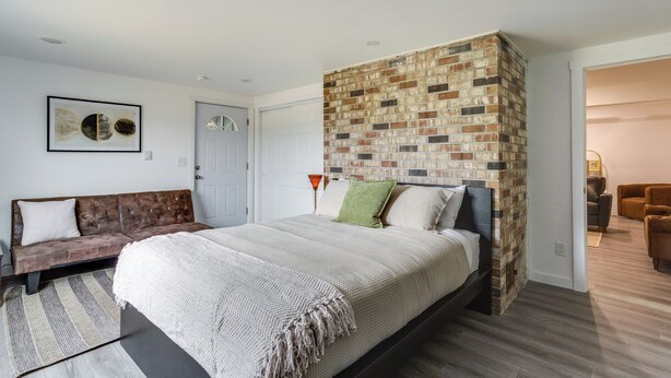 A bedroom with a brick accent wall, a bed with a white bedspread, a brown sofa, and a framed picture on the wall. - Quick Hwy Access to Seattle! Mtn-view Apt w/ Yard (Renton)