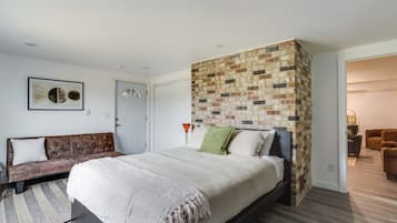 Um quarto com uma parede de destaque em tijolos, uma cama com colcha branca, um sofá marrom e um quadro na parede.