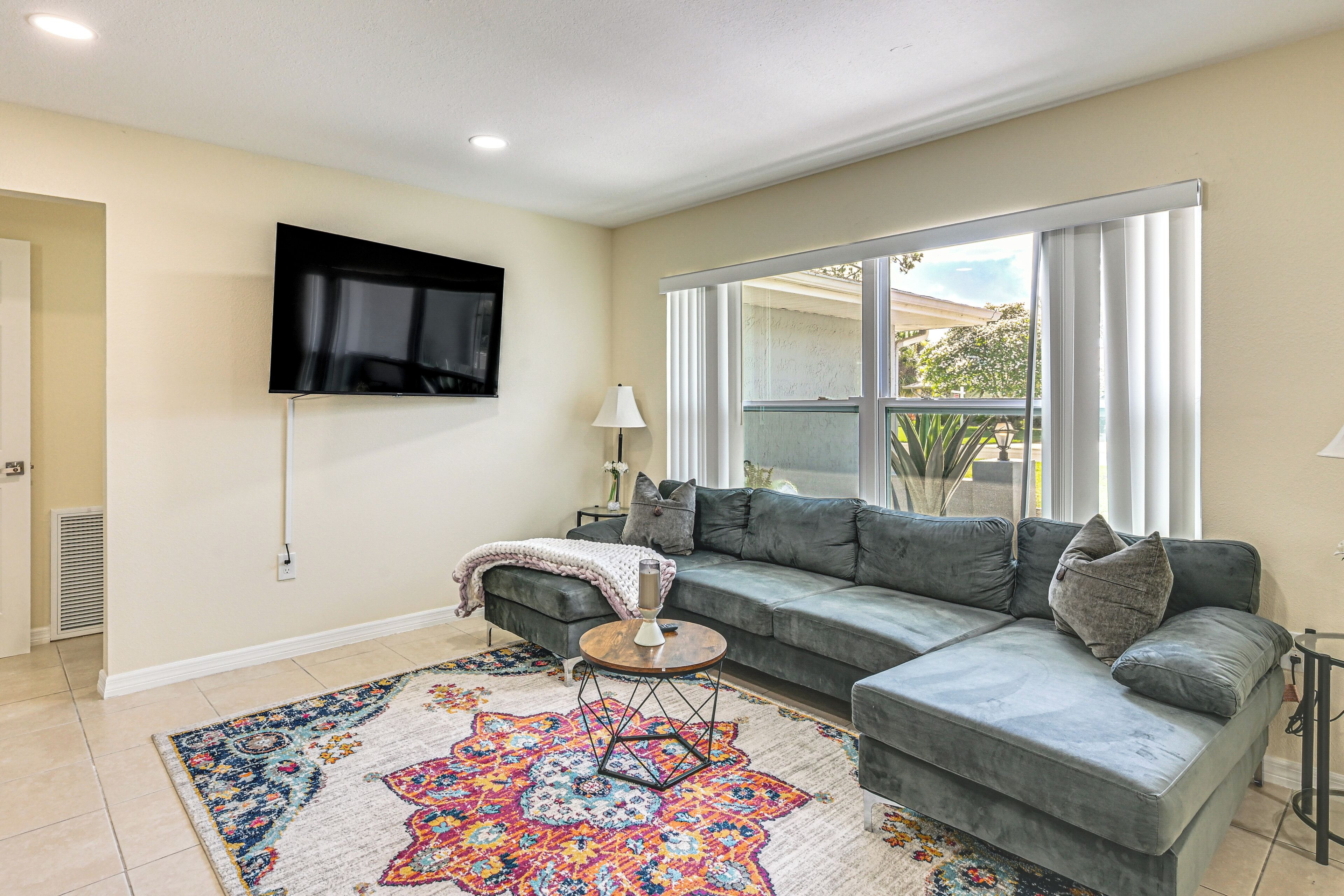A living room with a sectional sofa, a TV mounted on the wall, a patterned rug, and a window with curtains.