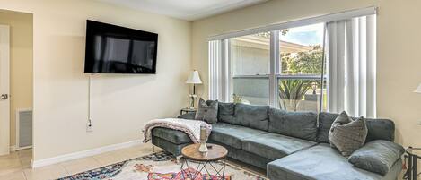 A living room with a sectional sofa, a TV mounted on the wall, a patterned rug, and a window with curtains.