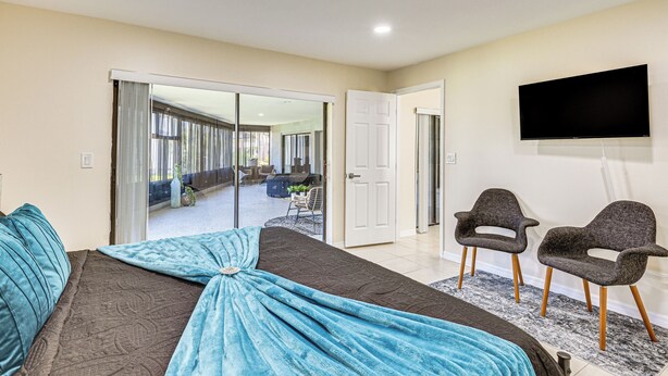 A bedroom with a bed, a TV, two chairs, and a sliding door leading to a balcony. - Enclosed Lanai w/ Hot Tub! New Port Richey Home (New Port Richey)
