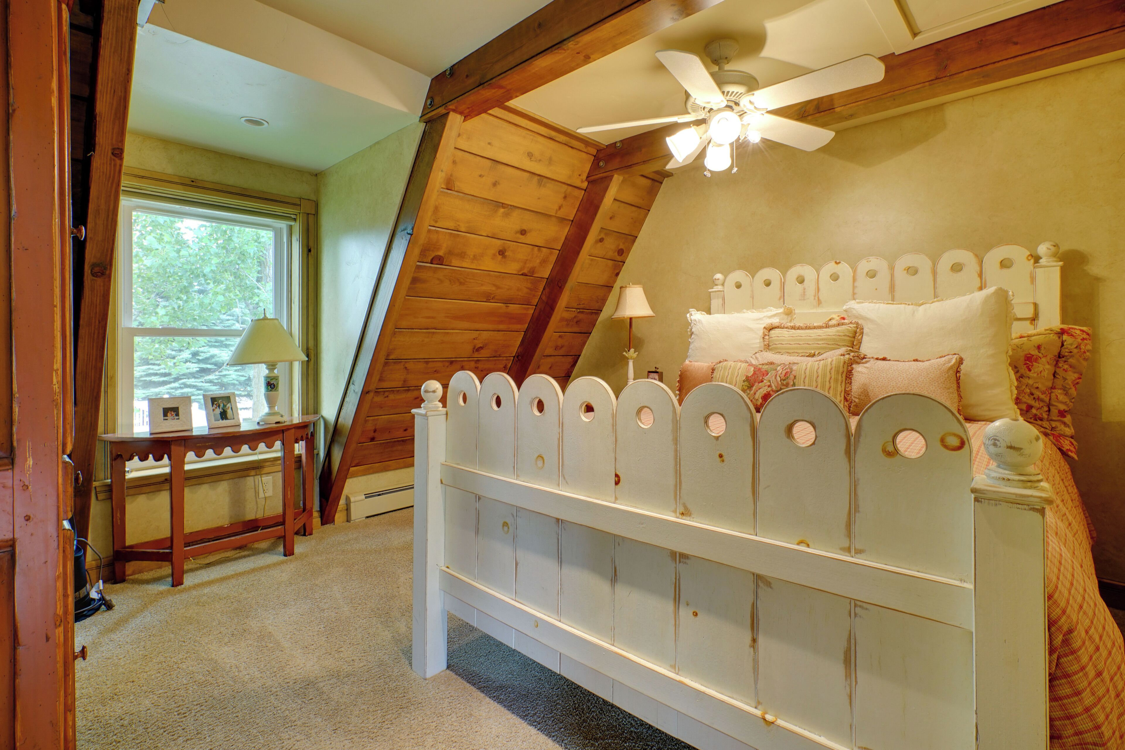 A bedroom with a sloped ceiling, a white bed with decorative headboard, a bedside table with a lamp, and a window with a view of trees.