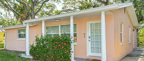 A single-story building with a porch, a small garden with red flowers, and a large tree in the background.