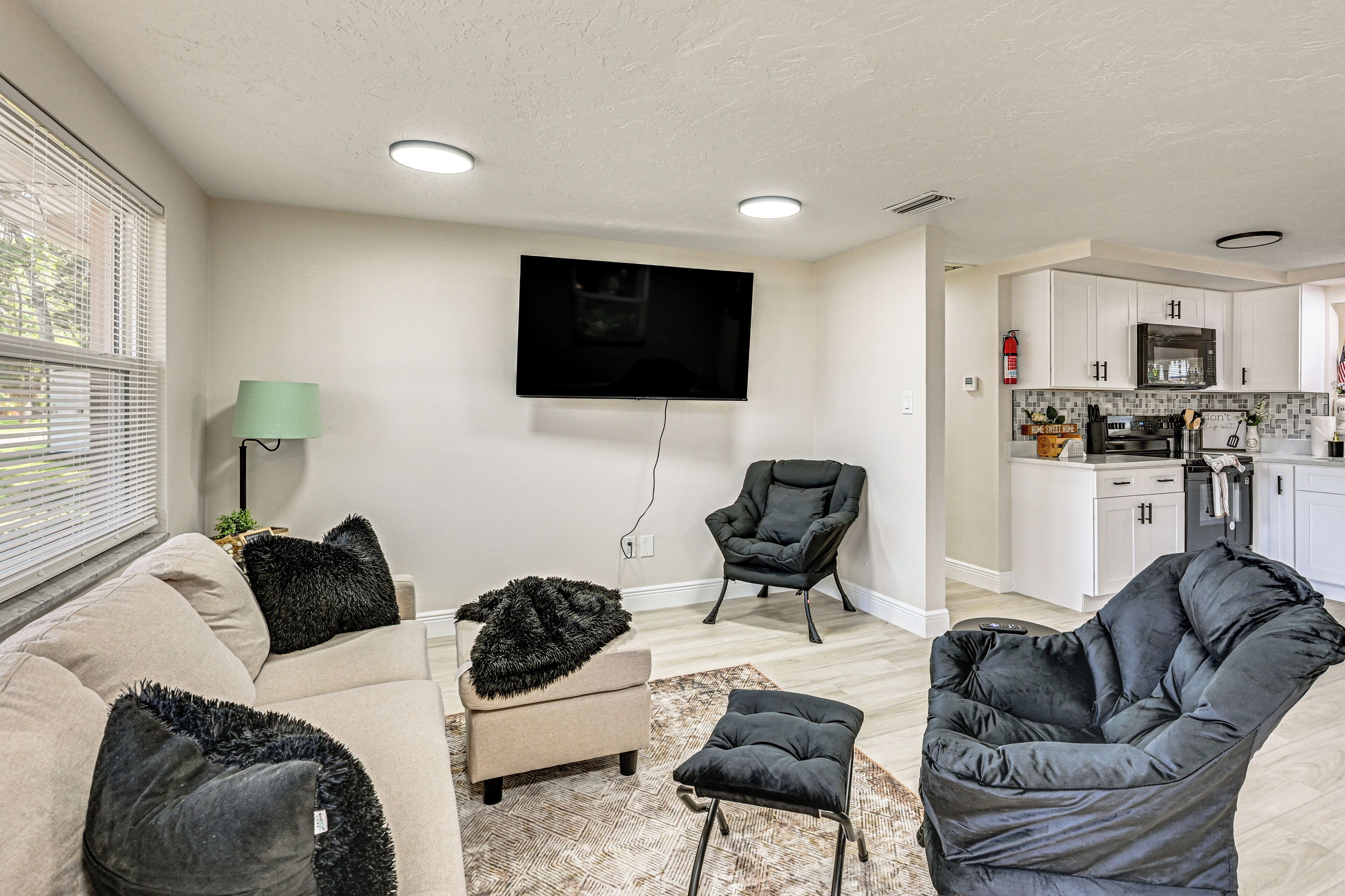 A living room with a sofa, ottoman, and two chairs, a flat-screen TV mounted on the wall, and a kitchen in the background.