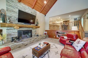 A spacious living room with a stone fireplace, red leather sofas, a wooden coffee table, and a TV mounted on the wall. - Hot Tub + Forest Views! Lake Arrowhead Cabin (Cedar Glen)