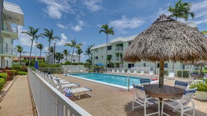 A pool area with lounge chairs, a thatched umbrella, and a view of a building with balconies and palm trees. - Steps to Pool & Ocean! Key Colony Beach Retreat (Key Colony Beach)