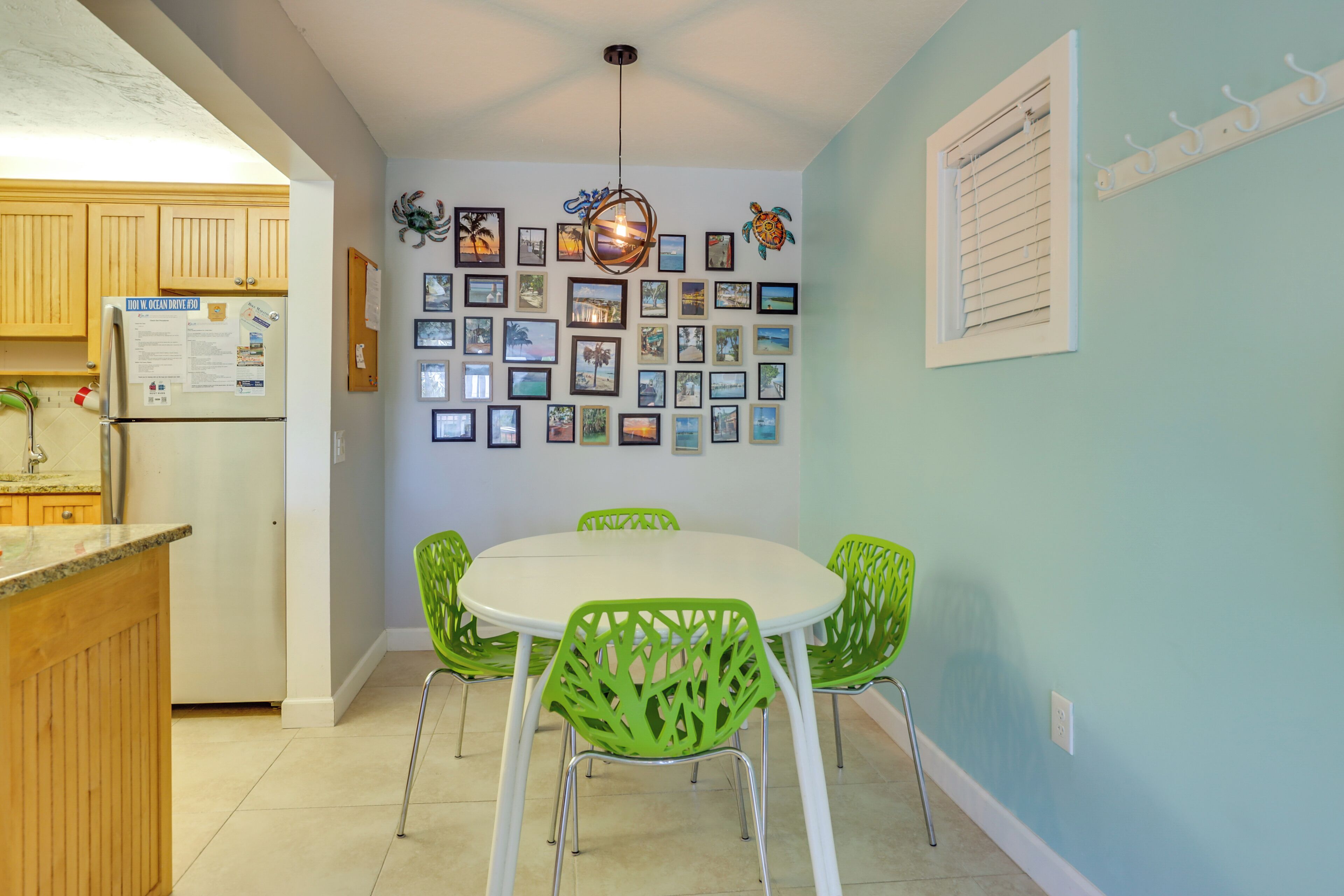 A kitchen with wooden cabinets, a refrigerator, a dining table with green chairs, and a wall decorated with framed photos.