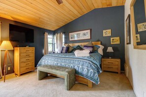 Un dormitorio con techo de madera, una cama con ropa de cama estampada, un banco, una mesita de noche y una lámpara.