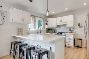 A modern kitchen with white cabinetry, a central island with bar stools, a microwave, and a refrigerator. - Spacious Kanab Basecamp Game Rm, Patio, Fire Pit (Kanab)