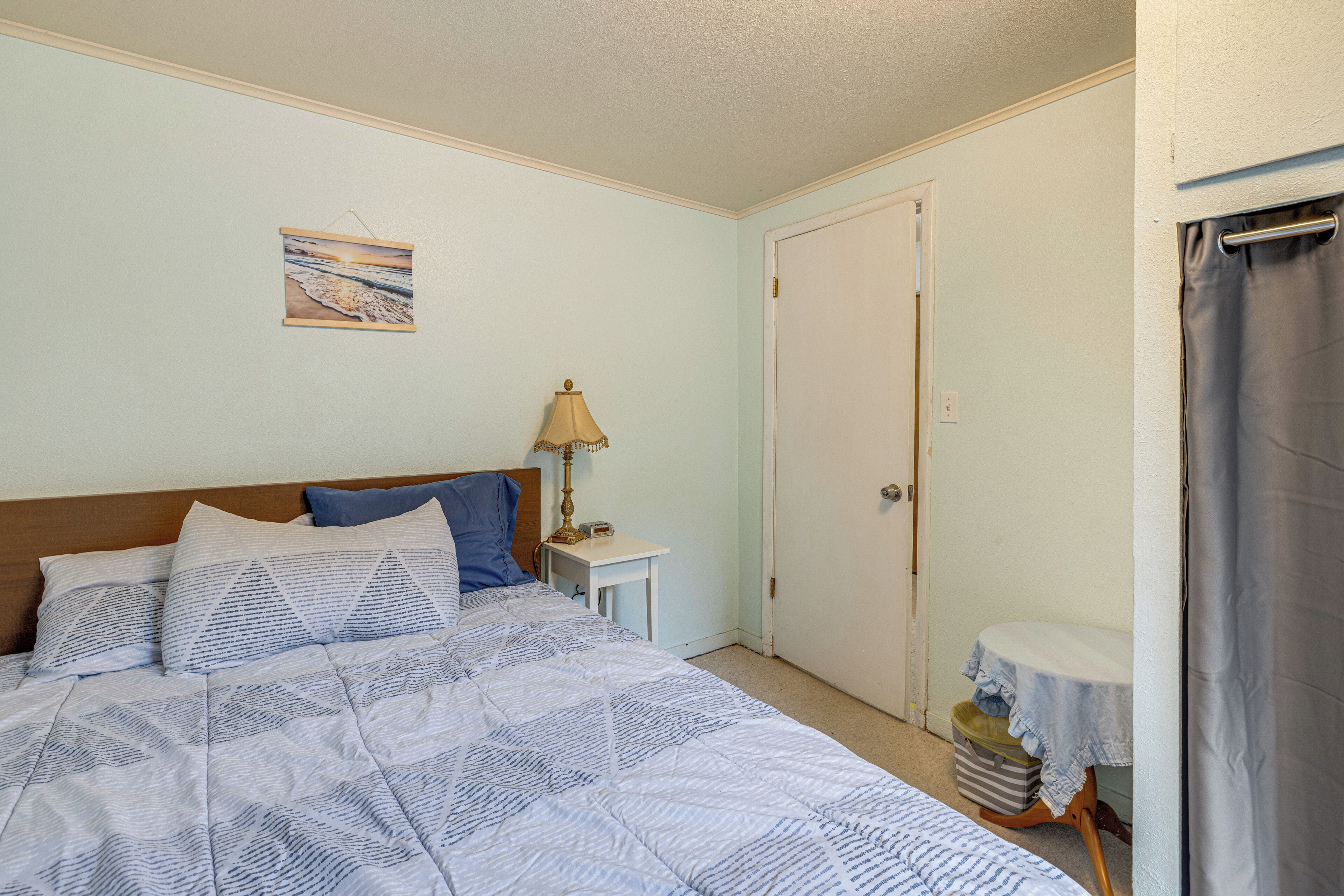 A bedroom with a bed, pillows, a nightstand, a lamp, a door, and a small table with a tablecloth.