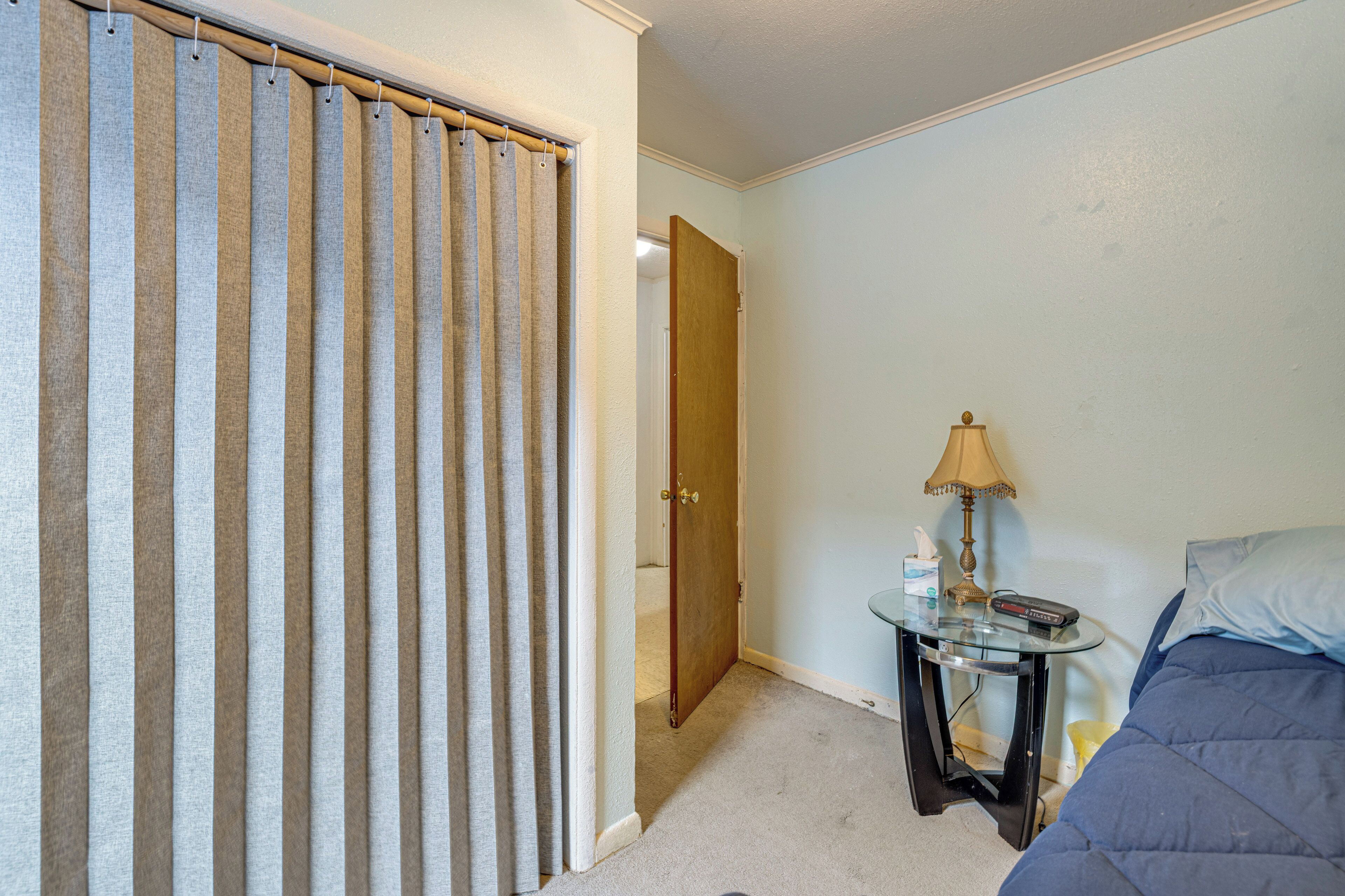 Een hotelkamer met een bed, een nachtkastje met een lamp, een kleintje tafeltje met een afstandsbediening, en een deur die leidt naar een andere kamer.