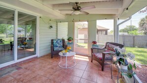 Une terrasse couverte avec un ventilateur de plafond, deux fauteuils en osier, une petite table agrémentée de fleurs et une vue sur le jardin arrière.