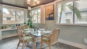Un coin repas avec une table en verre, des chaises en osier, un lustre et de grandes fenêtres offrant une vue sur les palmiers.