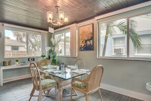 Un coin repas avec une table en verre, des chaises en osier, un lustre et de grandes fenêtres offrant une vue sur les palmiers.