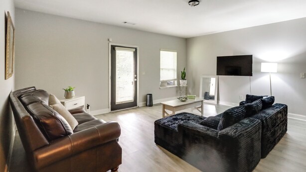Een woonkamer met een bruine leren bank, een zwart geweven poef, een witte bijzettafel met een plant en een televisie op een meubel.