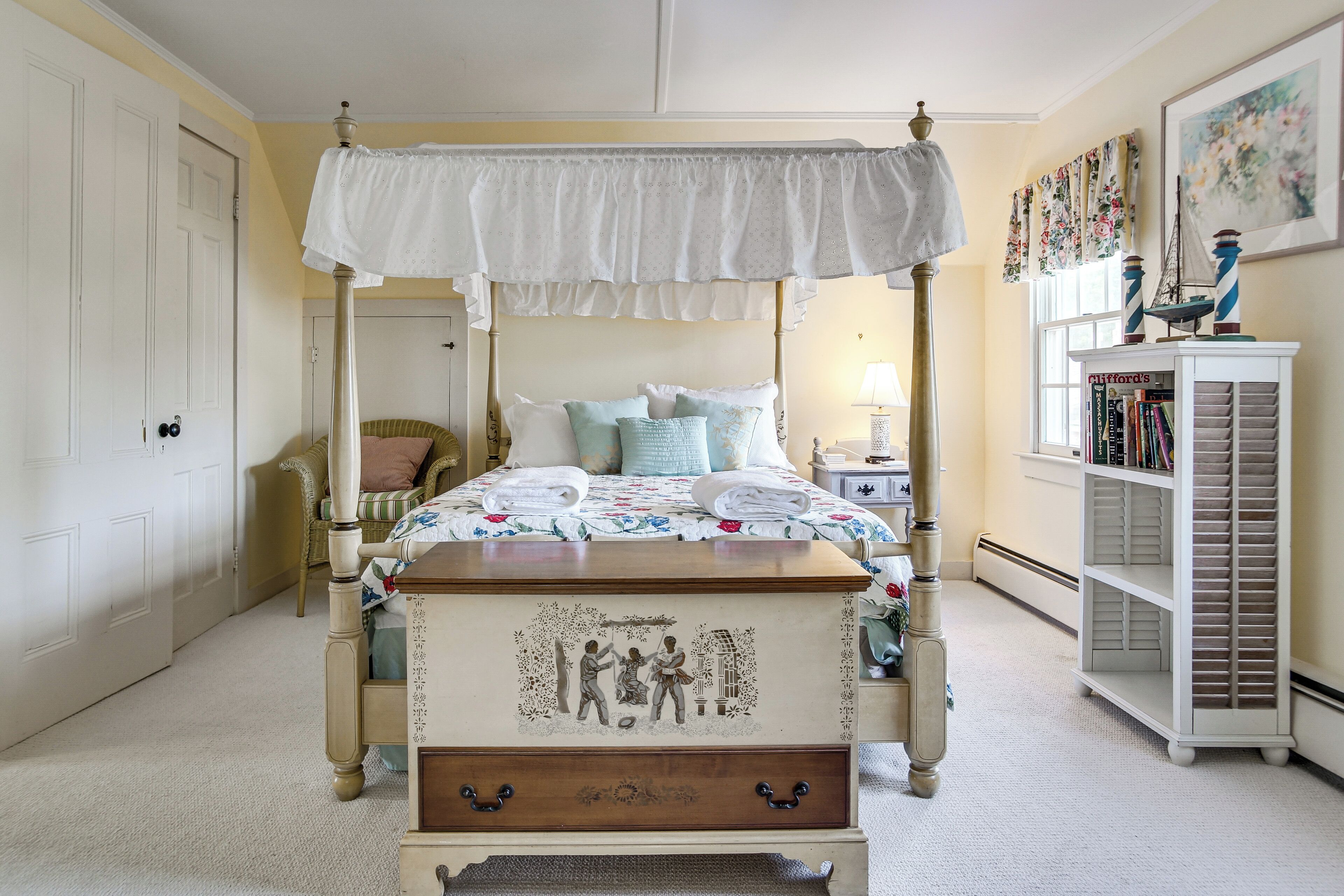 A four-poster canopy bed with a decorative headboard, a bedside table with a lamp, a bookshelf, and a chair in a room with cream walls and a carpeted floor.