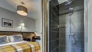 Ein Hotelzimmer mit Bett, Nachttisch, einer Wandleuchte, einem gerahmten Bild und einem Duschbereich mit Glastür.
